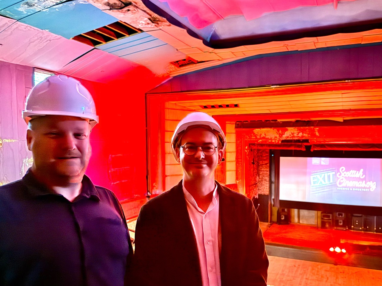 Gary Painter and Gordon Barr, wearing hard hats inside a historic cinema, with the ScottishCinemas logo projected onto the screen behind them.