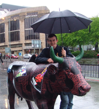 Gordon Barr holding a black umbrella behind the Chem-moo-stry cow sculpture, decorated with scientific patterns and green horns.